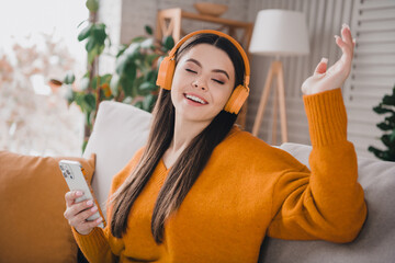 Young woman enjoying music with orange headphones while seated comfortably on a couch in a cozy...