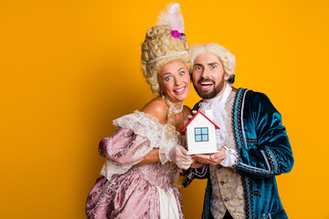 A Joyful Couple in Elegant Historic Costumes Holding a Small House Model Against a Vibrant Yellow...