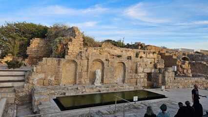 Obraz premium Ancient Roman bath ruins at Caesarea Maritima, Israel, with a reflecting pool and visitors exploring the site.