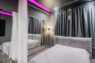 Modern bedroom with a large bed, dark walls, and a mirrored wall. White sheer curtains and purple accent lighting add to the room unique ambiance