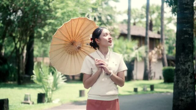 Young Thai woman wearing traditional Lanna dress, showcasing elegant cultural attire while standing with decorative parasol amid lush tropical garden surroundings