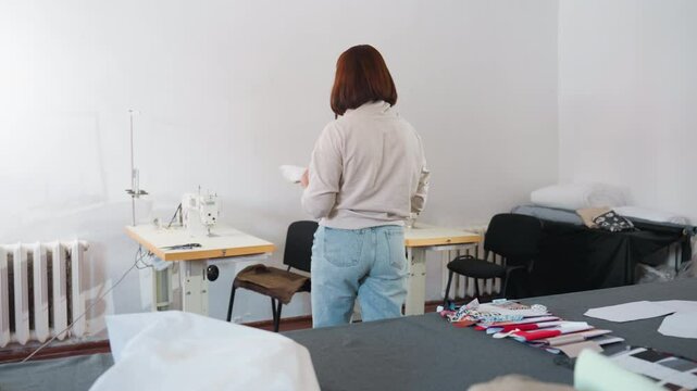 Young lady holding sewing thread preparing to work in sewing workshop with industrial sewing machine, fabric pieces, sewing materials scattered on large table, ready for garment creation