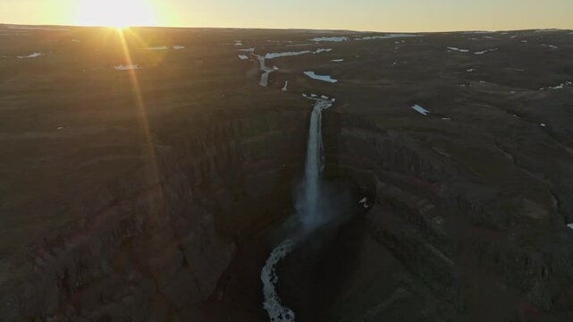 Aerial view of Hengifoss waterfall in East Iceland, captured at sunset with dramatic light rays and deep canyon shadows highlighting the basaltic rock formations.