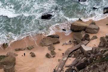 A sunny beach scene with golden sand, gentle waves and a rocky cliff topped with houses under a vibrant blue sky in Nazaré, Portugal.
