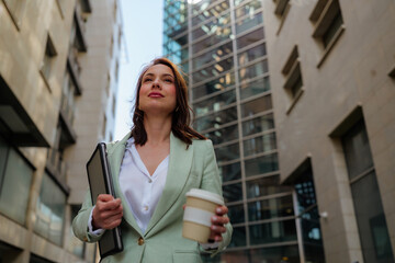 Confident businesswoman walking down the street, holding a laptop and a coffee, looking up at...