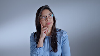 Woman in glasses and blue shirt, hand on chin, thoughtful pose, gray background, close-up shot,...