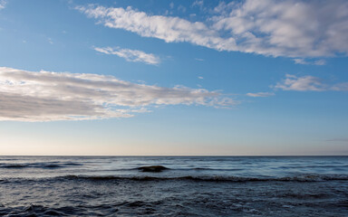 Calm sea with small waves under a blue sky with scattered clouds at twilight. A dark shadow from a wave is visible on the water's surface due to the side and slightly backlighting.