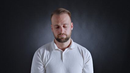 Fototapeta premium A man in a white shirt stands with eyes closed, conveying a sense of contemplation or exhaustion. The dark background accentuates his introspective mood.