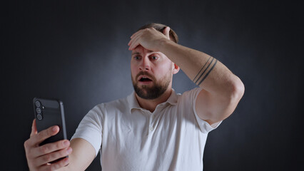 Man in white shirt holding phone, forehead on hand, looking shocked, tattoo on arm, dark background.