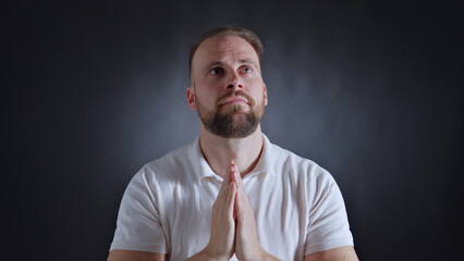 A man clasps his hands in prayer, gazing upward with a solemn expression, conveying a sense of hope...