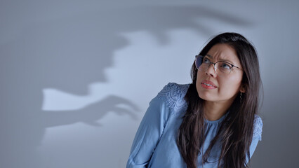 Woman in glasses looks puzzled, shadow of a hand claw behind her, close-up shot, gray background.