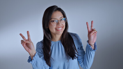A woman in a blue shirt and glasses flashes a peace sign, exuding positivity and joy. The close-up...