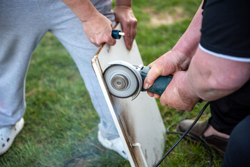 Man using angle grinder to cut wooden board with help outdoors on grass lawn.