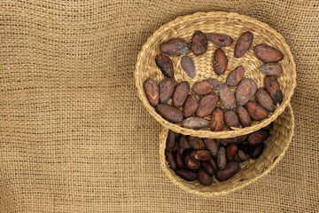 Cocoa beans in woven basket placed on a textured burlap fabric background. Ideal for...