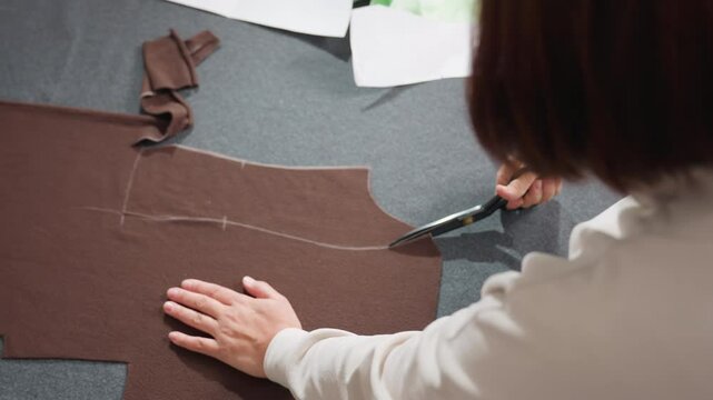 Woman placing hands on material cutting precisely along pattern lines on brown fabric using sharp scissors while creating garment pieces, surrounded by scattered paper patterns and fabrics