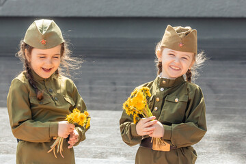 Patriotic education of children. Little girls with long braids in a military uniform, military cap, hold flowers in hands against the background of the memorial. Photo for the Victory Day on May 9