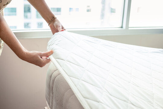 hands of woman making bed, tucking in mattress pad and linens in lighted room with large window. Hotel room cleaning