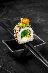 Chopsticks holding uramaki sushi roll with sesame seeds and vegetables over soy sauce