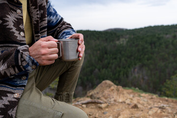 Close-up of hiker's hands holding a metal cup with a hot drink, enjoying a moment of peace in a...