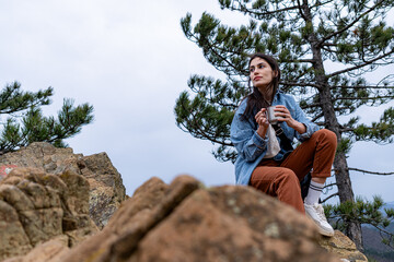 Female hiker taking a break and drinking a hot beverage from a metal cup while sitting on a rock on top of a mountain