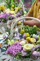 Close-up of a male florist in a yellow T-shirt holding a large Easter basket with lilac and yellow flowers and bunnies