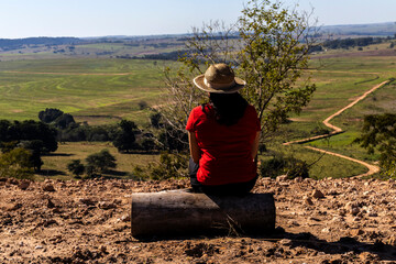 Woman sitting on a wooden stump looks out over the landscape of rural roads and farmland from a mountain range in Brazil