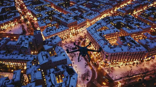 Drone is flying over a city at night. The city is lit up with lights and the drone is flying over a snowy street