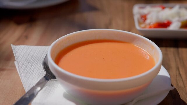 Traditional Spanish cold soup, Gazpacho, served in a white bowl with a spoon on a napkin, accompanied by a tapa, on a wooden table
