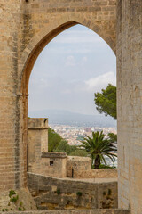 Looking through and ancient stone arch there is a view of the city of Palma in the background, a palm tree in mid ground with blue sky in Mallorca Spain.