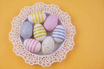 A white lace doily basket filled with colorful, striped, and speckled Easter eggs sits against a solid yellow background