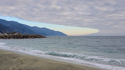 Waves gently roll onto a sandy beach of the Cinque Terre with a rocky jetty and distant mountains under a dramatic, cloud-layered sky.