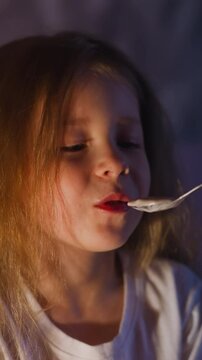 Smiling little blonde girl grudgingly eats porridge while mother feeds before going to sleep in semidark bedroom at gentle night lamp light slow motion