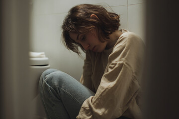 Fototapeta premium Young woman sits on floor, head bowed, wearing beige sweater and jeans, in a dimly lit bathroom, conveying sadness and solitude