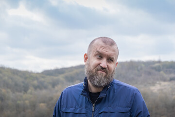 Man with beard expresses uncertainty outdoors in a forested area under cloudy skies, showcasing...