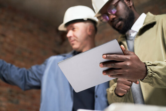 Low angle shot of silver tablet in hand of African American interior designer standing near Caucasian foreman