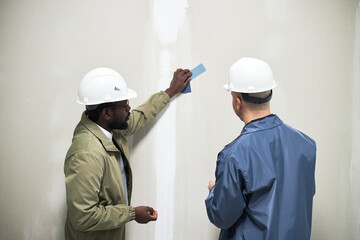 Two male biracial interior designers wearing plastic hardhats while discussing wall color of color...