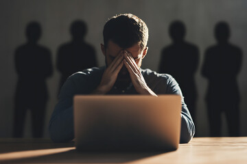 Frustrated man in front of laptop with head in hands. The background consists of four shadowy figures which could indicate external pressures on the individual.