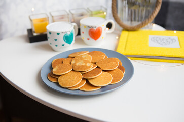 A plate of mini pancakes sits on a white table alongside mugs and candles, creating a cozy breakfast scene