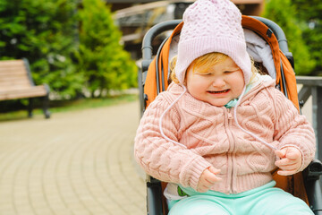 A young child in a cozy sweater and hat sits in a stroller while crying in a vibrant park Lush trees and pathways surround the area providing a lively atmosphere for outdoor activities