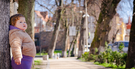 A young child stands beside a tree in a park taking in the warmth of a sunny day The setting...