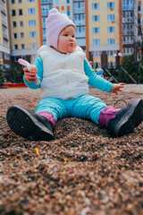 Child enjoying time in a sandbox at an urban playground wearing a warm vest and pink hat covered in...
