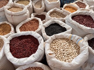 Top view of various types of beans, lentils, and grains in large white fabric sacks arranged closely together at an outdoor local market. Natural dry food assortment for sale.