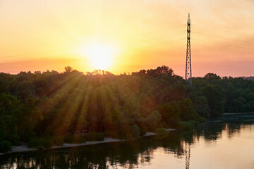 Golden sunrise over a calm river and lush green forest with a silhouette of a power line tower.