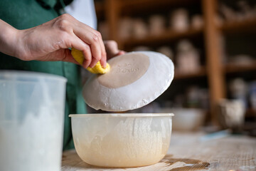 Potter covers ceramic saucer with glaze using sponge and jug in workshop, applying final touch. Artistic pottery expertise, ceramic clay glazing, handmade craftsmanship, ware smooth finish and care