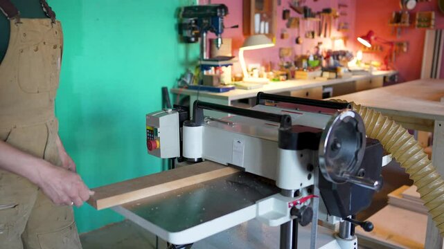 Jointer machine in wood shop provides smooth planing, preventing chips on knots against grain. Unrecognizable joiner woodworker work on final board thickness and high quality wood finish, manual labor