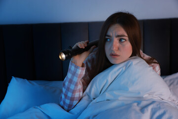 Fear of darkness. Scared young woman with flashlight lying on bed in room at night