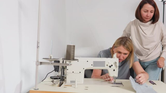 Fashion designers adjusting needle on bar tack sewing machine, one helps partner then returns to seat, showing synergy, professional environment, shared skill, efficient production