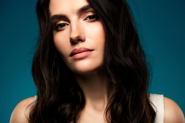 Stylish portrait of a young brunette woman with natural makeup and glowing skin on a blue gradient...