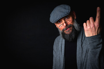 Elderly bearded man in cap and glasses shows a finger up gesture, expressing sarcasm or rebellion....