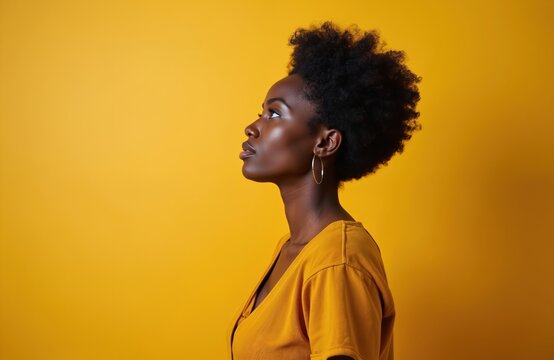 Side profile of black woman with natural afro hair. Wearing gold hoop earrings and yellow blouse, she looks up with focused expression. Set against yellow backdrop.